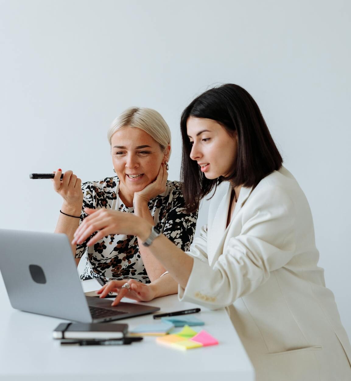 Two women sit at a desk working together on a laptop, using Smartsheet to organize tasks, with pens, a smartphone, and colorful sticky notes nearby.