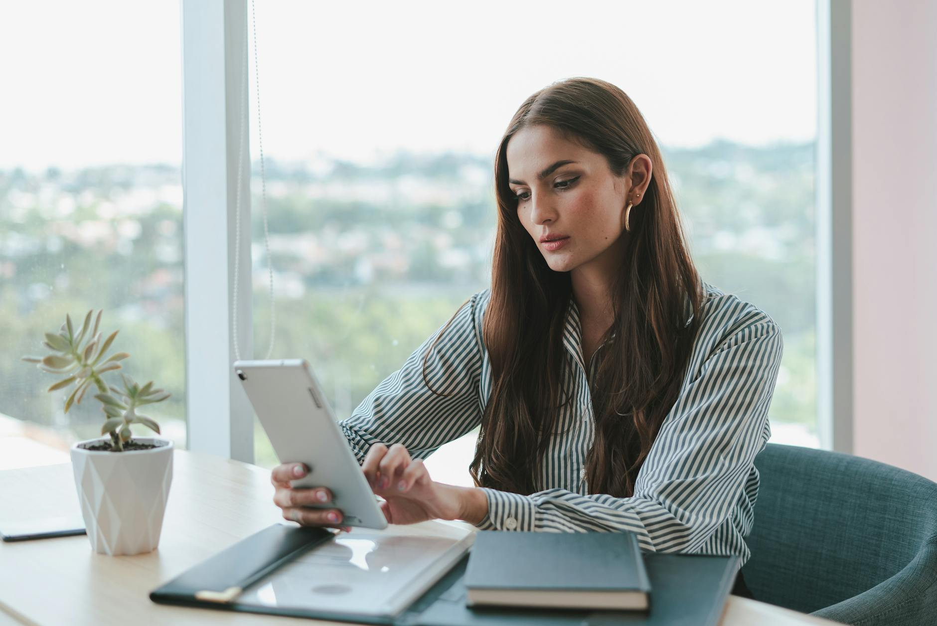 A woman sits at a desk by a window, using a tablet with Smartsheet. There are notebooks and a small potted plant on the desk.
